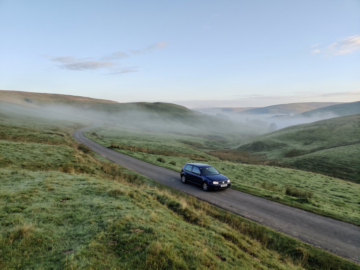 Ein kleines blaues Auto fährt auf einer Landstraße durch eine Hügellandschaft im Morgennebel